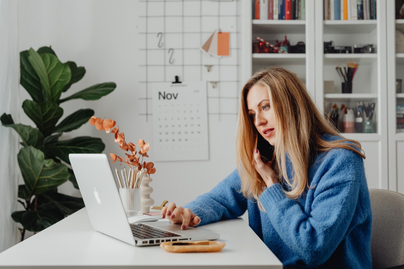 A woman on a phone call at a tidy home-office desk, a wall calendar behind her